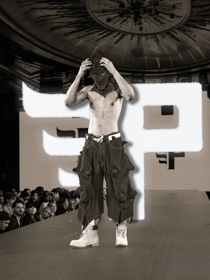 Model in a black hood and cargo trousers walking a runway under a domed ceiling