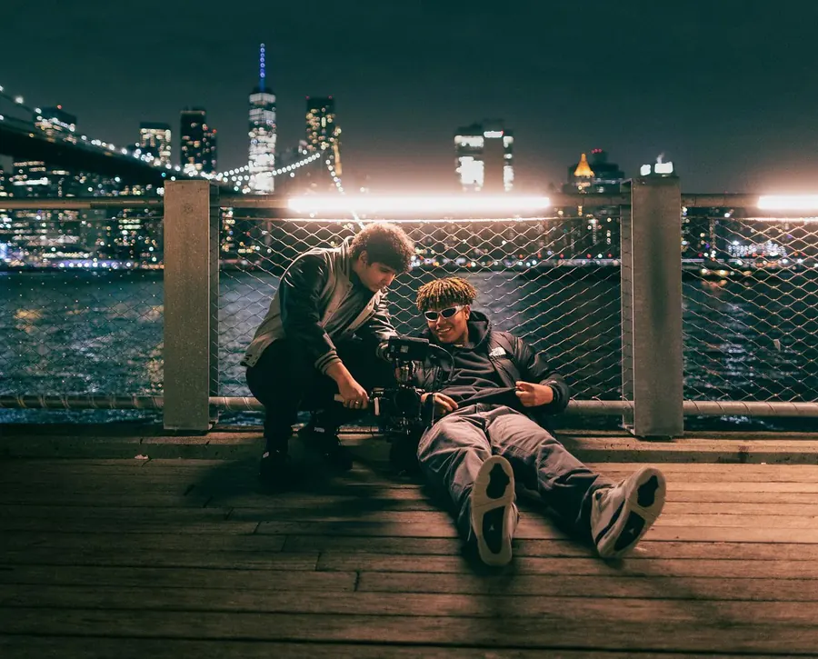 Two men resting on a pier across from the Manhattan skyline at night